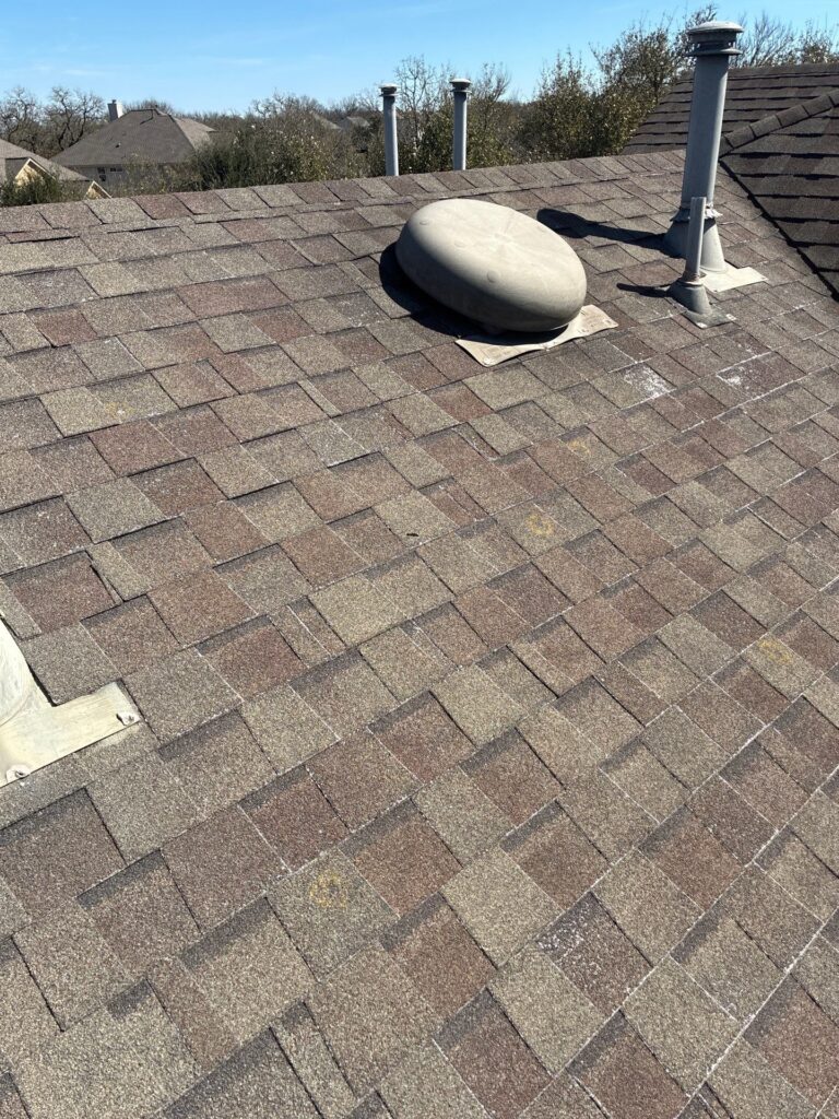 Rooftop with brown shingles, featuring a circular skylight dome and several metal vent pipes nearby under a clear blue sky.