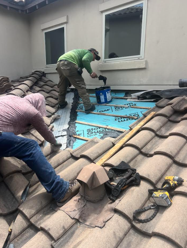 Two construction workers install blue underlayment on a tiled roof near a dormer window; one kneels with a tool bucket nearby.
