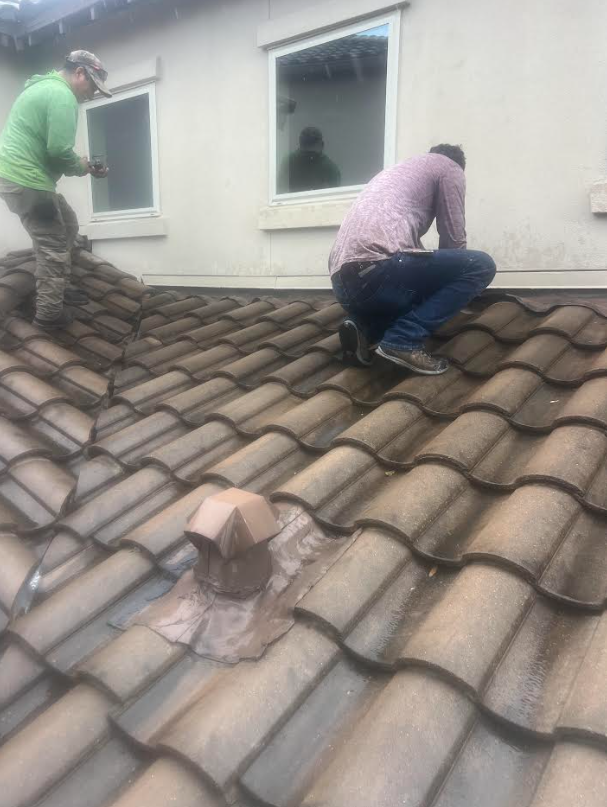 Two workers on a tiled roof patching around a vent near a beige house, one in a green shirt and cap, the other kneeling in a pink shirt.