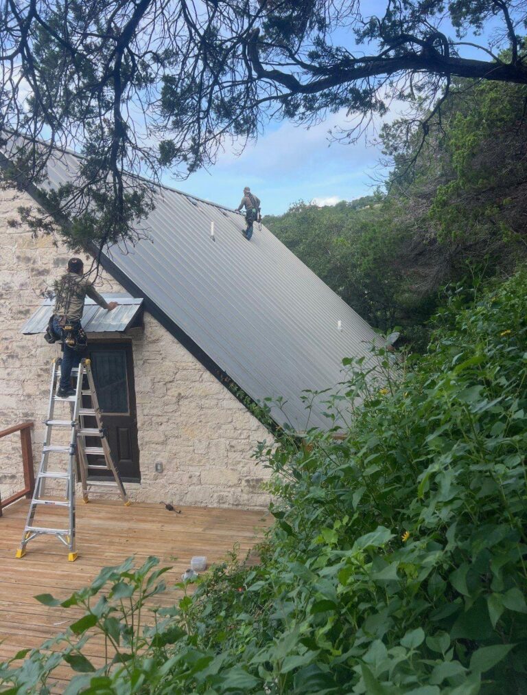 Two workers on a sloped metal roof; one stands on a ladder at a stone house and another up higher in safety gear, surrounded by greenery.