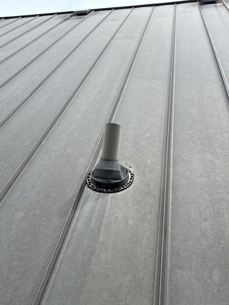Close-up of a gray standing-seam metal roof with a round vent pipe and flashing at the center bottom.