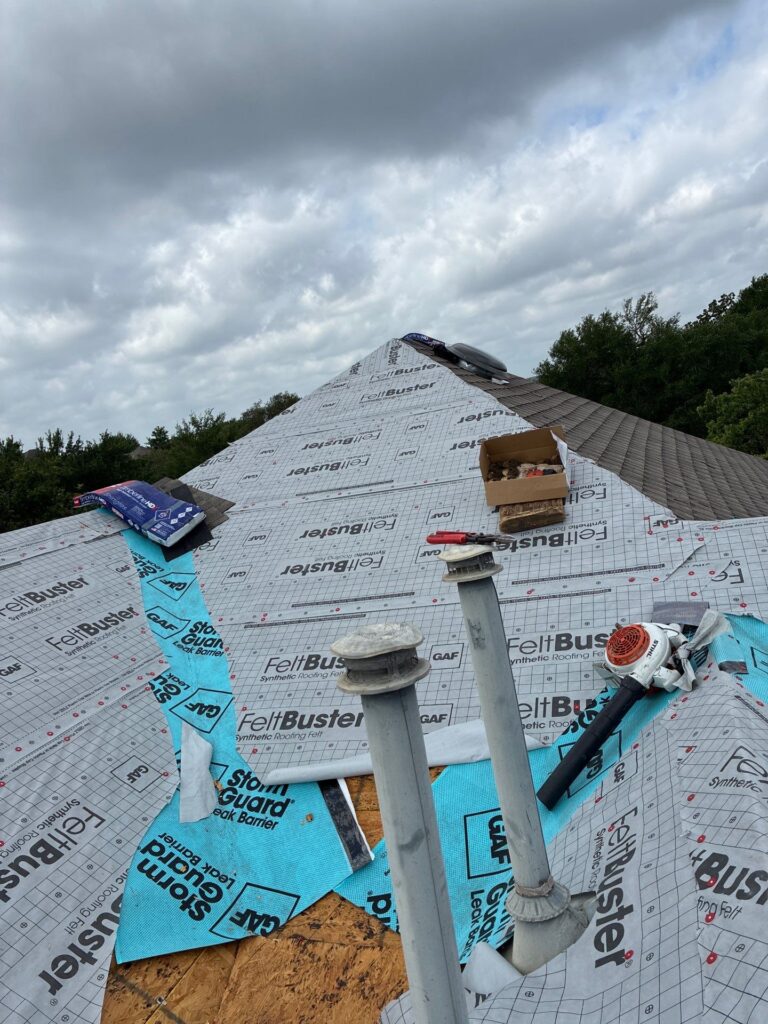 Rooftop under construction with weatherproof FeltBuster underlayment, vent pipes, and a toolbox on the sloped roof amid cloudy skies.