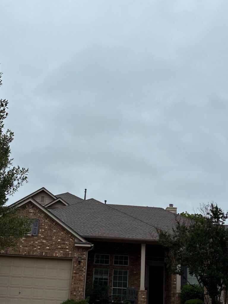 Front view of a brick suburban house with a two-car garage, a gray shingle roof, and some trees against a cloudy sky