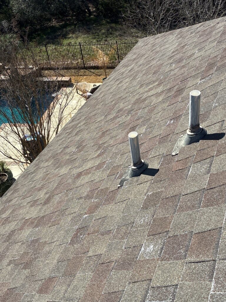Roof with three white vent pipes protruding from gray shingle surface; backyard with fence and trees in background.