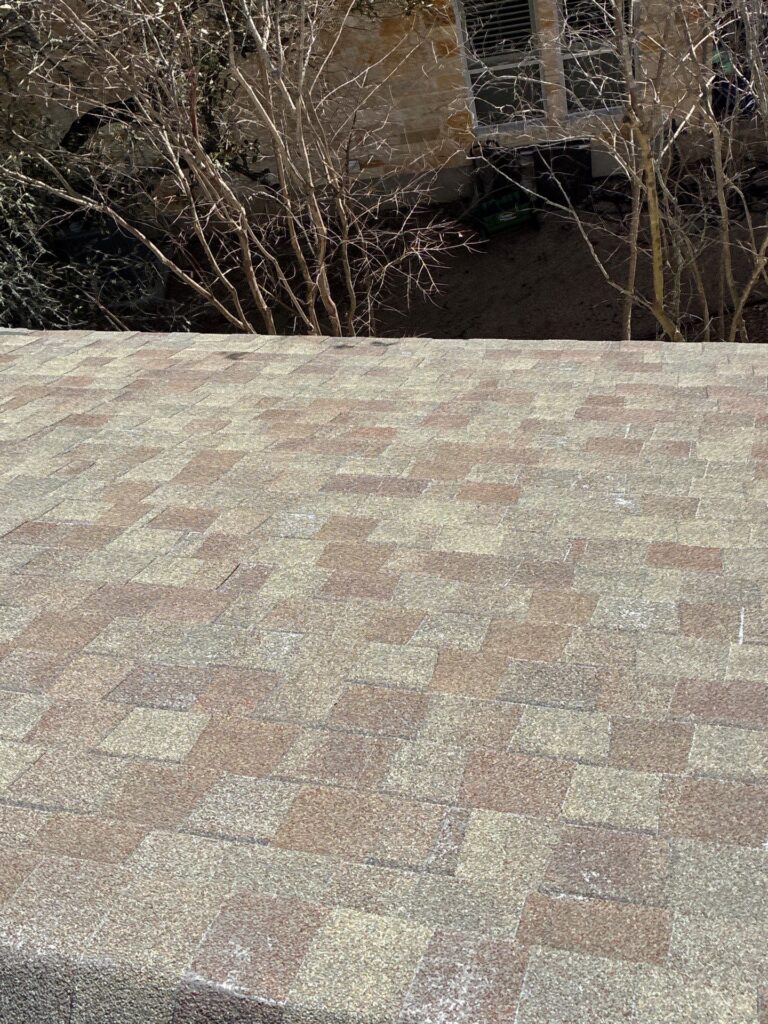 Close-up of a brown tiled roof edge with a yard and bare trees behind a stone wall and window.