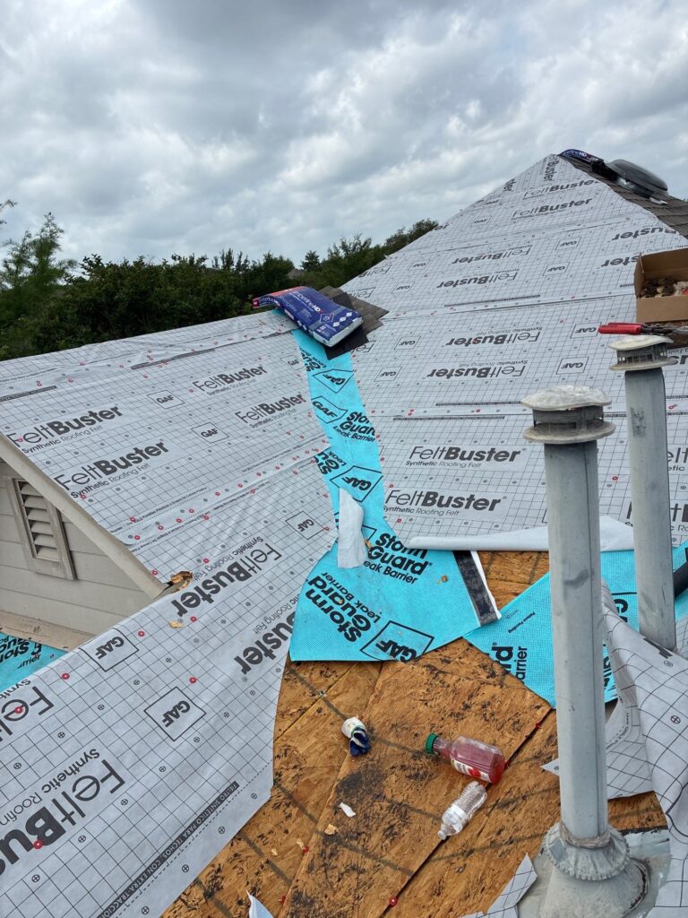 Roof under construction with FeltBuster roofing underlayment, blue protective strip, gloves, and scattered tools on plywood sheathing under a cloudy sky.