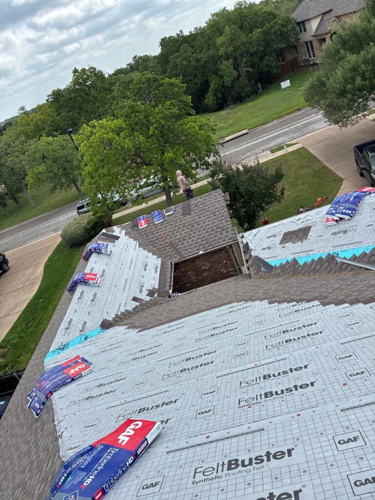 Worker in a hoodie stands on a residential roof under construction, with FeltBuster roof underlayment and packaging nearby.