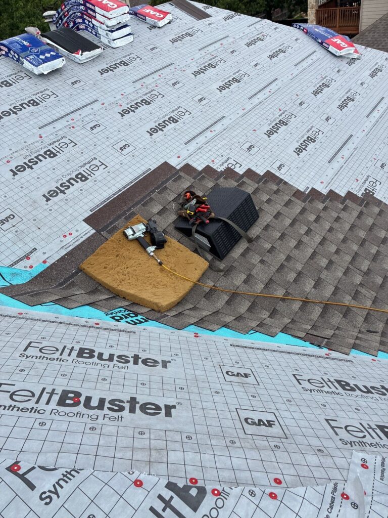 Roof underlayment being installed with bundles of insulation along edges and roofing tools on asphalt shingles.