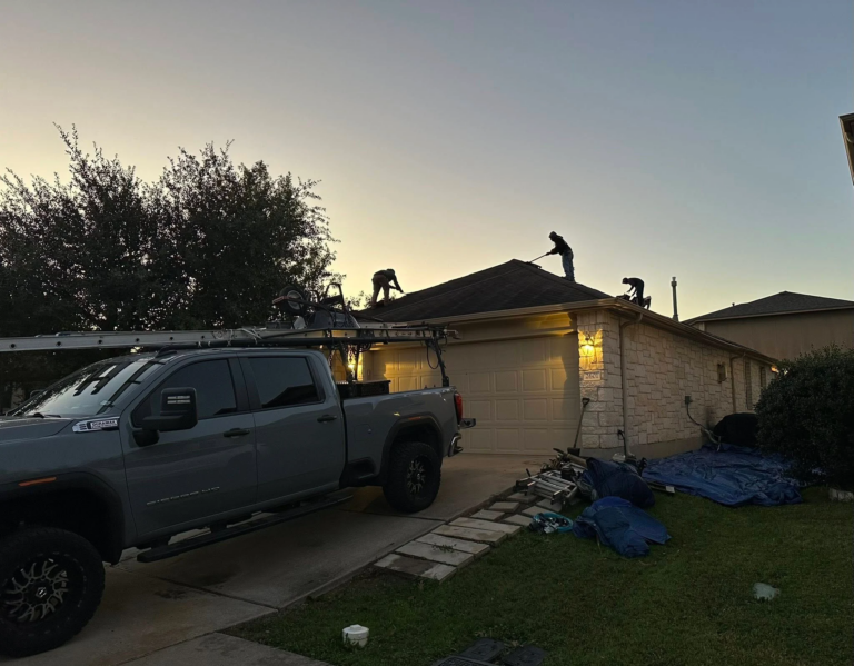 Three workers on a sloped roof performing roof work at sunset, with a ladder truck parked in the driveway nearby.
