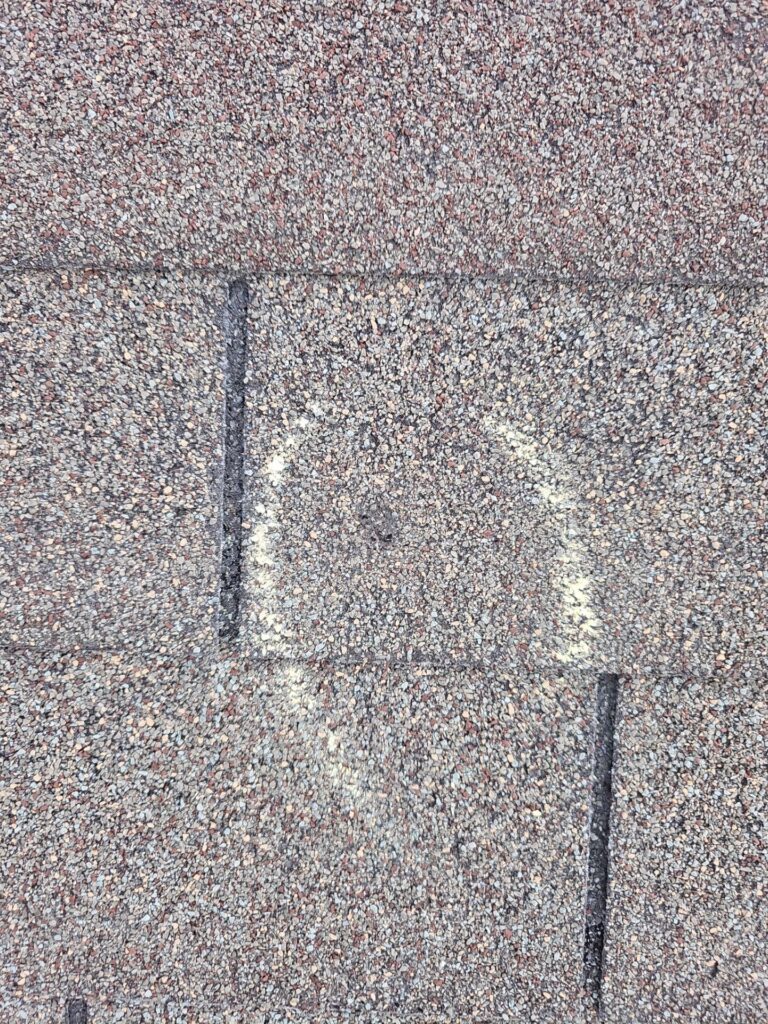 Close-up of an asphalt roof shingle with a rough granulated surface and a dark vertical seam, showing weathering patches.