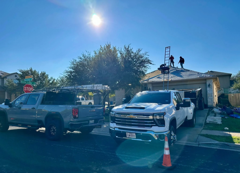 Two workers on a house roof with a tall ladder, bright sun overhead, and parked trucks nearby in a driveway.