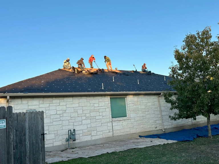 Five workers on a pitched roof replacing shingles, clear blue sky above and a tree to the right.