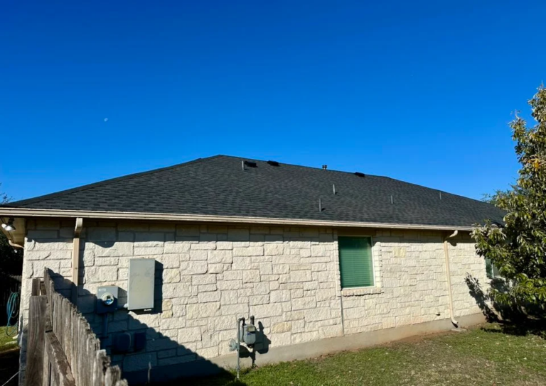 Side view of a single-story stone house with a dark shingled roof, green shuttered window, and a wooden fence against a clear blue sky.