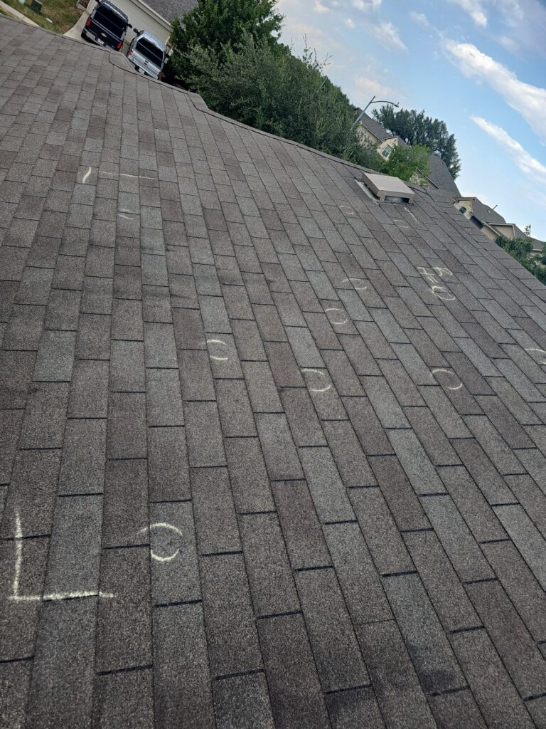 Pitched asphalt shingle roof with chalk inspection marks scattered across many shingles; a small rooftop vent is visible, with a suburban street and trees in the background under a blue sky