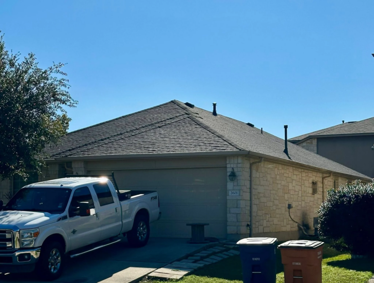 White pickup truck parked in a suburban driveway beside a brick house under a clear blue sky, with several trash bins in front yard.