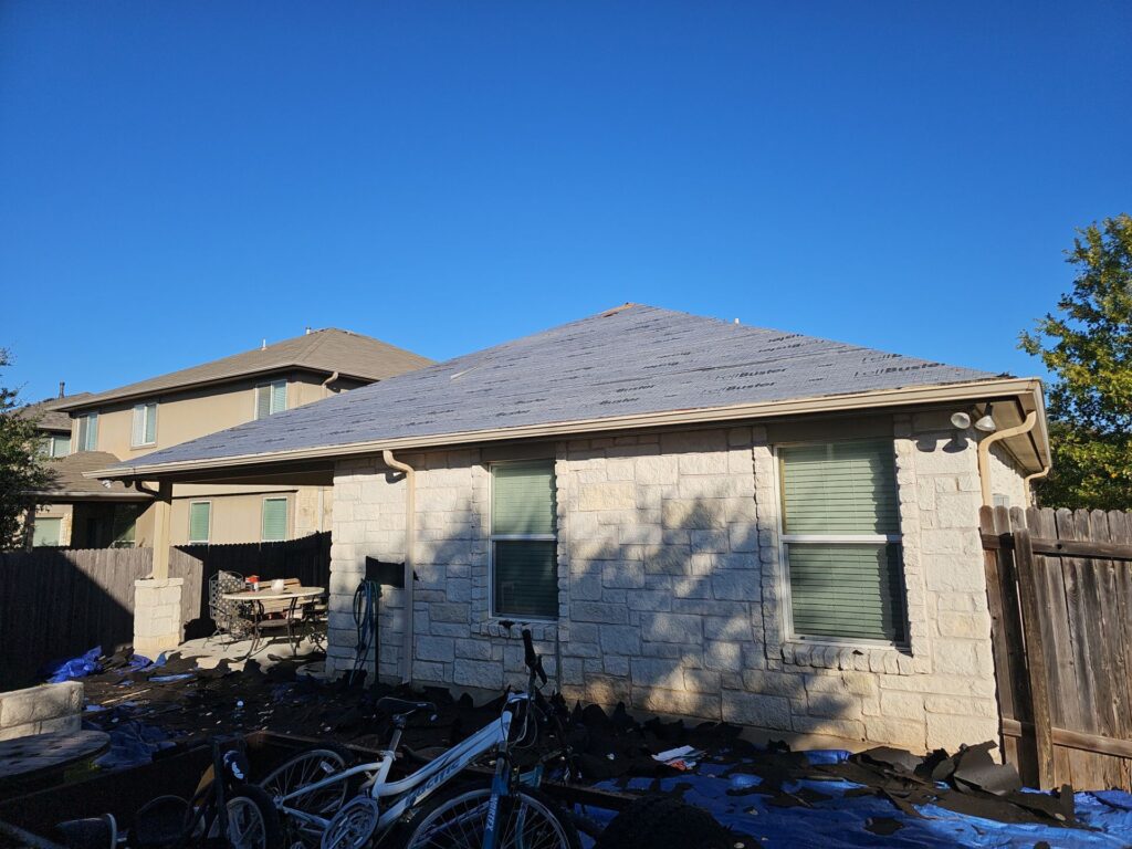 Backyard of a brick house with a partially stripped roof and construction debris on the ground, including bicycles and blue tarps, under a clear blue sky.