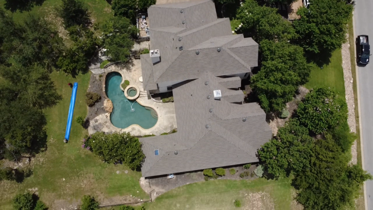 Aerial view of a large house with a kidney-shaped pool and attached spa, surrounded by trees and a blue water slide on the lawn to the left.