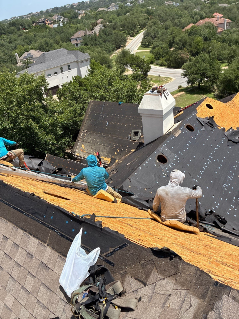 Construction workers on a sloped roof removing old shingles, with tools and tar paper visible