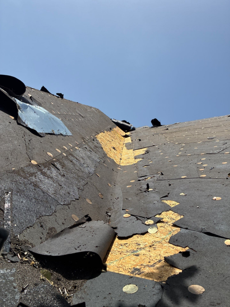 Damaged roof with torn black roofing membrane, exposed plywood, and scattered debris under a clear blue sky.