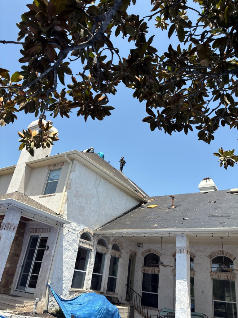 Construction workers on a roof above a white stucco house, blue tarp in the yard, clear blue sky overhead.