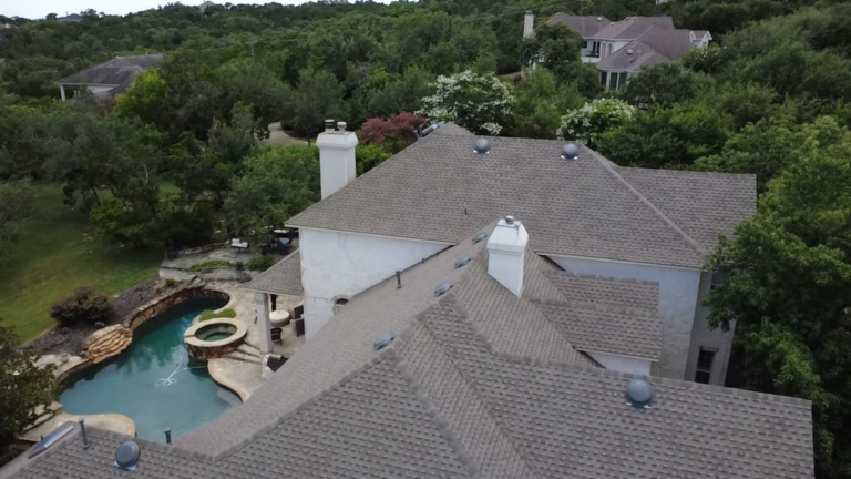 Aerial view of a large white house with gray tiled roof, lush trees, and a curved backyard pool with a spa.
