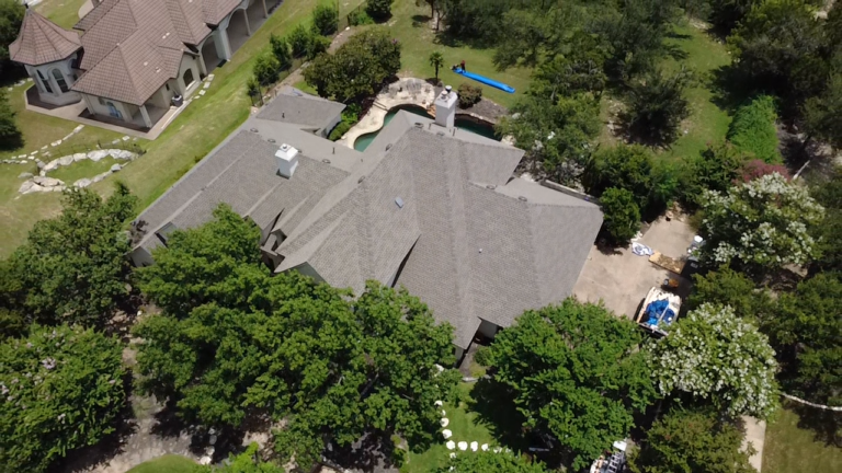 Aerial view of a large gray-roofed suburban house with a backyard pool and surrounding trees.