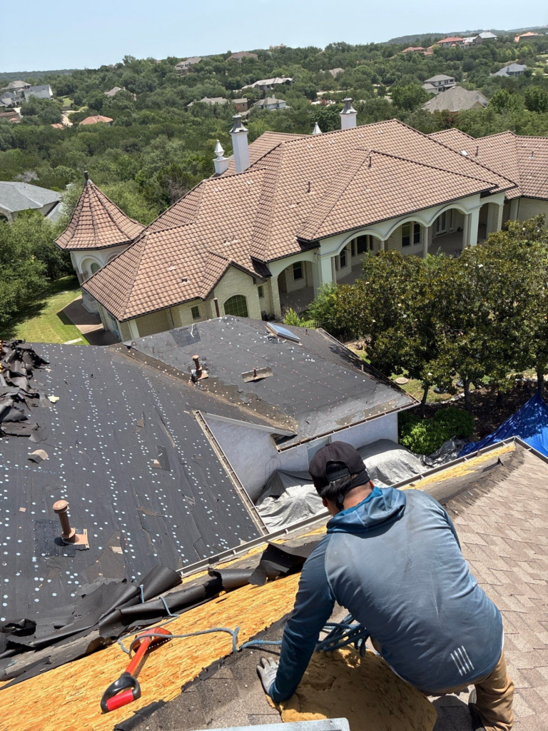 Construction worker on a sloped roof removing old shingles with a toolbox and debris nearby, overlooking upscale houses and trees.