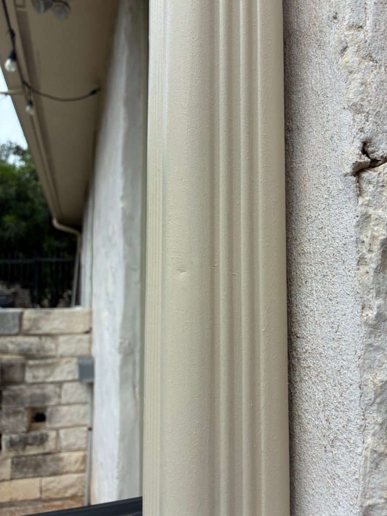 Close-up of a beige fluted column beside a textured stone wall, with a blurred outdoor background.
