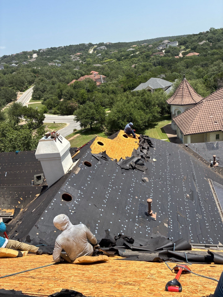 Workers repair a damaged black roof with torn shingles on a sunny hillside house backyard.