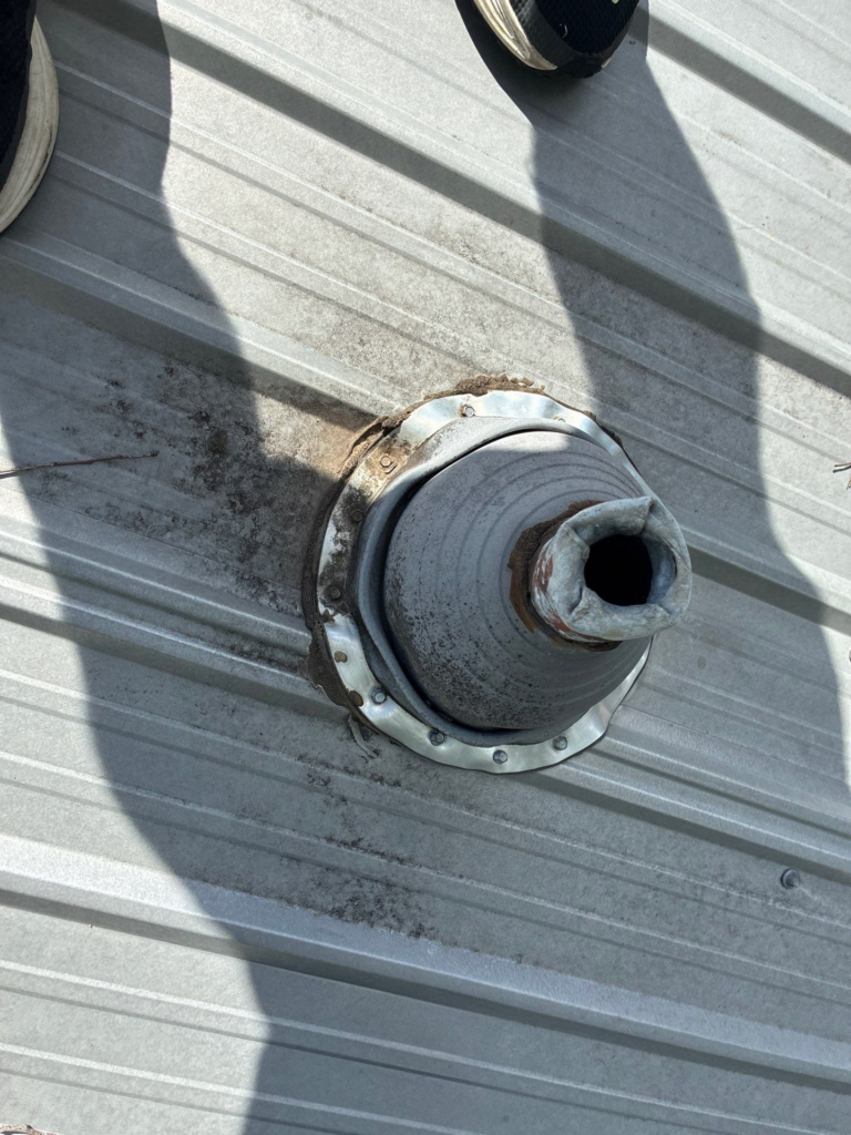 Close-up of a metal roof vent flashing with a rubber boot, showing rust and dirt around the flange on a corrugated roof.