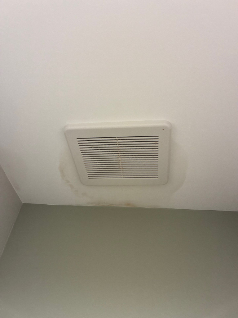 White ceiling vent with a rectangular grille mounted near a corner, with light brown stains around its edge.