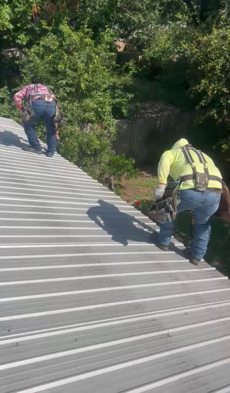 Two construction workers in safety harnesses on a slanted metal roof amid trees and a yard. Pulling or fastening metal panels while wearing tool belts.