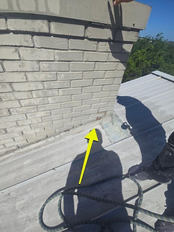 On a flat metal roof beside a brick chimney, a yellow arrow points to a seam or flashing along the roof edge, with a rope and a worker's shadow nearby.