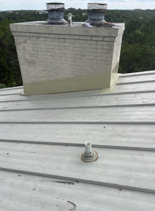 Rooftop view of a white brick chimney with two vent caps; a white vent pipe rises from the metal roof in the foreground, with trees in the distance.