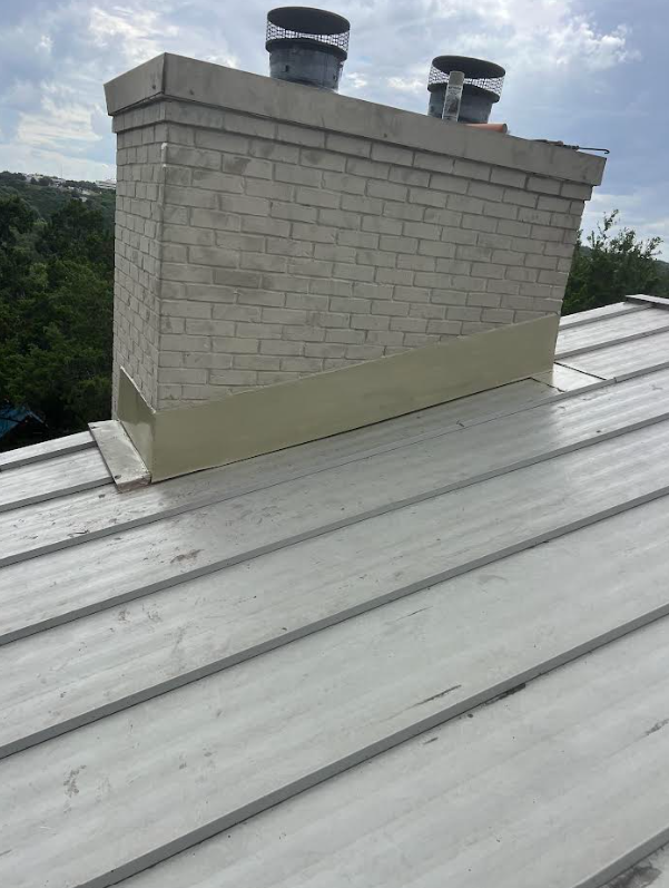 Metal standing-seam roof with a brick chimney structure and two circular vent pipes on top, under a cloudy sky