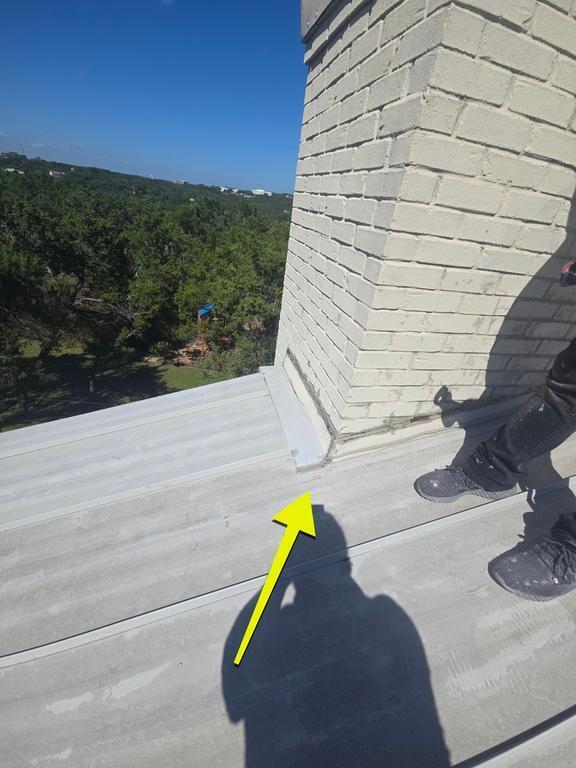 Arrow points to a seam/gap between metal roof panels beside a white brick chimney, with a worker’s boots and shadow visible on the roof and a blue sky backdrop.