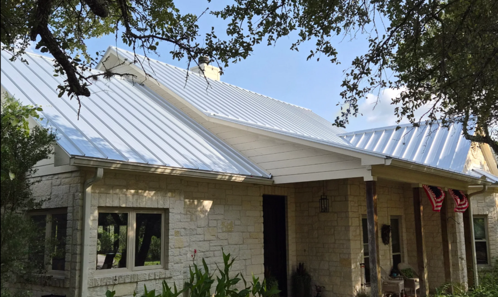 Front view of a light stone house with a white metal roof, a covered porch, and American flags hanging from the right side of the porch.
