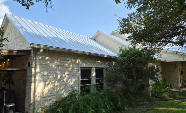 Screenshot 2026-07-10 143456 - Driftwood Builders Roofing Stone house with a light metal roof, downspout visible, and lush greenery in the front yard under a clear blue sky