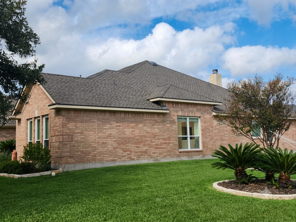 Single-story brick house with a gray shingled roof, white-framed windows, and a manicured lawn with a circular plant bed in the foreground.