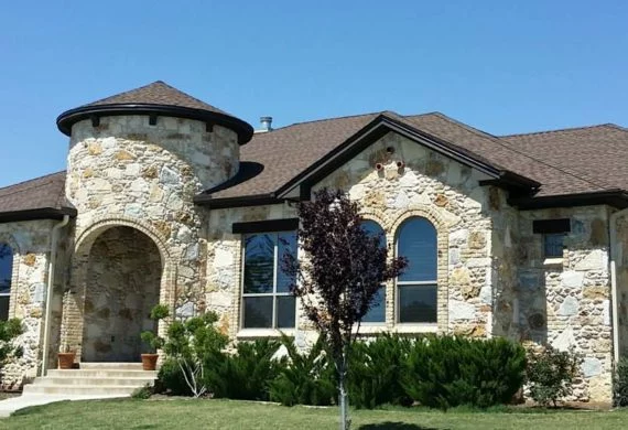 Stone residence with a turret, arched entry, and tan stone exterior; front lawn with shrubs and a small tree.