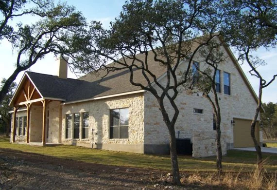Stone house with a wooden front porch, beige stone walls, and trees in the yard on a sunny day.