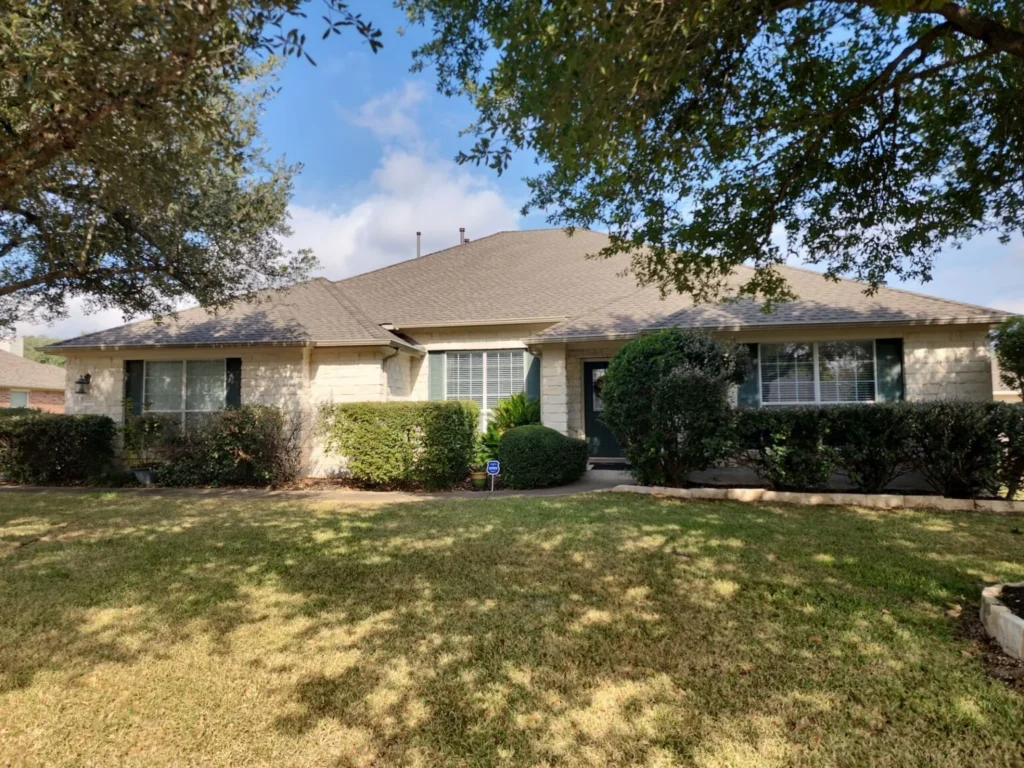 Single-story beige brick house with a well-kept lawn and trimmed hedges, framed by large trees above.