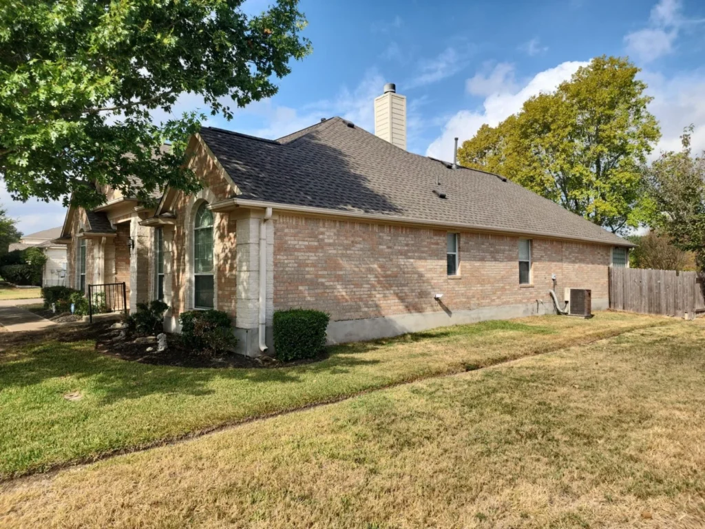 Side view of a single-story brick house with a brown shingle roof, arched window, and white trim, bordered by a lawn and shrubs on a sunny day.