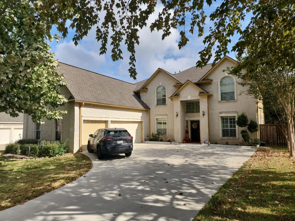 Front view of a two-story beige brick house with arched windows, a two-car garage, and a black SUV in the driveway, framed by trees.