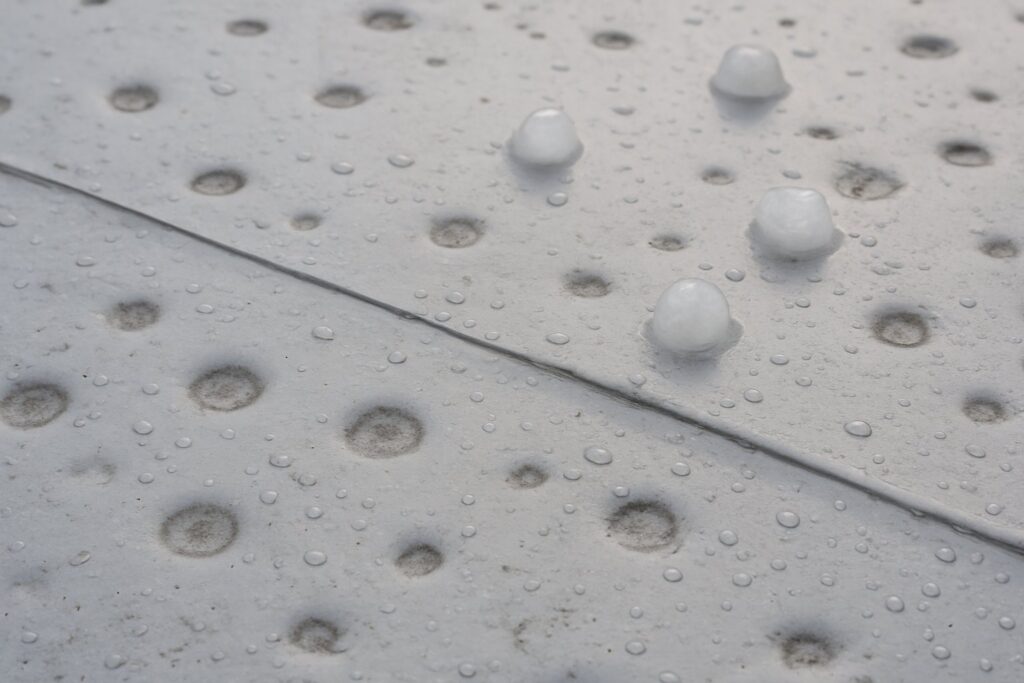 Close-up of a light gray metal surface covered with many circular dimples and scattered droplets, with a diagonal seam running across the panel.