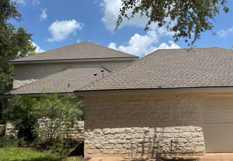 Beige stone house with intersecting tan shingle roofs, a closed garage door, and green shrubs against a bright blue sky with clouds.