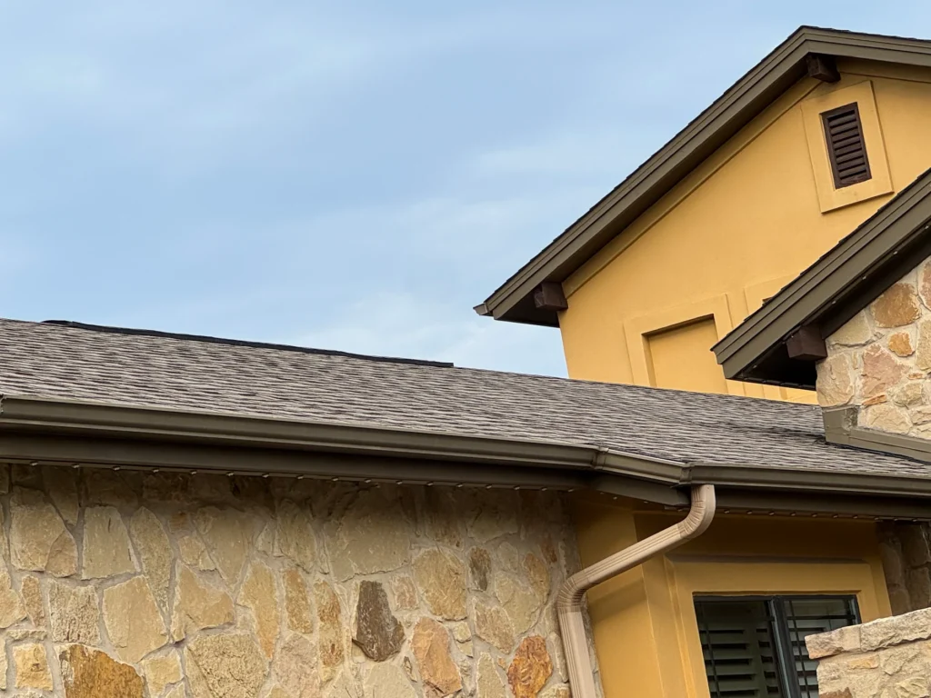 Exterior view of a stone-and-plaster house with tan walls, a sloped shingled roof, and a beige curved downspout clip running along the wall.