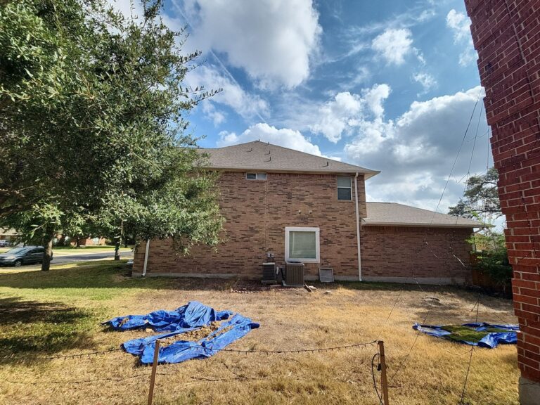 Backyard with a brick house, large tree on the left, and blue tarps covering dirt piles on dry grass under a sunny sky.