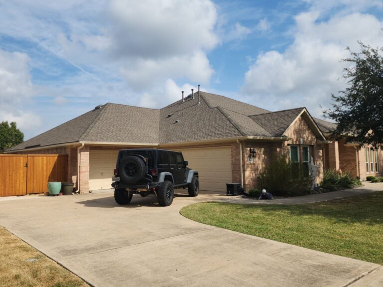 Black Jeep Wrangler with spare tire on a driveway in front of a beige brick, two-car garage suburban home under a blue sky with scattered clouds.