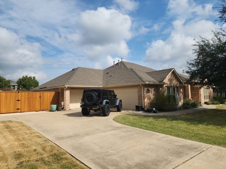 Black Jeep Wrangler parked in the driveway of a single-story brick house with beige garage doors and a wooden fence on the left, under a partly cloudy blue sky.